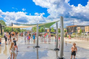 Kids playing in the splash pad. The sky is blue with puffy clouds. 